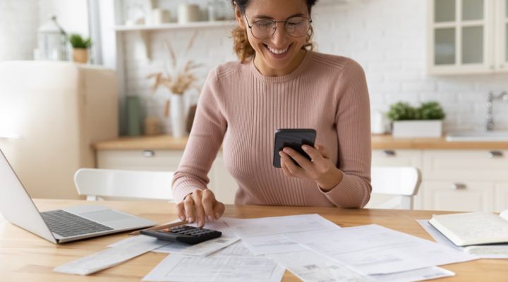 Smiling young 30s woman in eyewear looking at smartphone screen, feeling satisfied with fast secure online service, paying household bills taxes or insurance, managing budget, calculating expenses.
